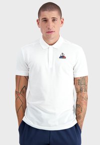 Polo shirt blanc en tissu respirant avec un col à boutons. Il présente un logo coloré sur la poitrine gauche et des manches courtes.