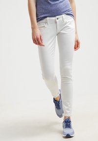 Jeans blancos ajustados con una textura suave y diseño de cinco bolsillos, combinados con zapatillas deportivas azules que cuentan con detalles de malla y cordones.