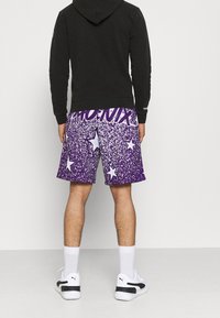Shorts violet à motif tacheté avec des étoiles blanches, assortis à un sweat à capuche noir. Des chaussettes blanches et des baskets noires et blanches complètent le look.