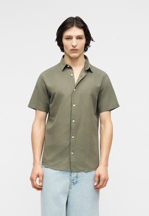 Νέος άνδρας με μεσαίου μήκους σκούρα μαλλιά, ντυμένος με κοντομάνικο πουκάμισο χακί και ανοιχτόχρωμο μπλε τζιν, στέκεται μπροστά σε απλό φόντο.