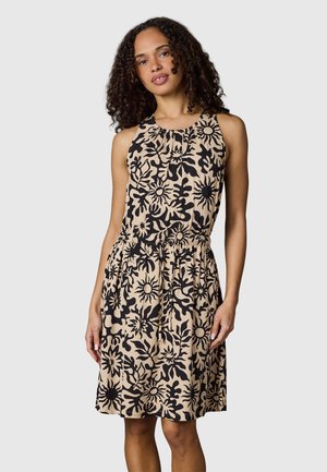 Kobieta o kręconych włosach, ubrana w beżową, bezrękawnikową sukienkę z czarnymi abstrakcyjnymi wzorami słonecznymi i kwiatowymi, stojąca na tle jednolitego jasnego tła.