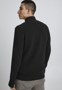 Schwarzer gerippter Pullover mit hohem Kragen, langen Ärmeln und strukturiertem Muster, getragen mit hellgrauen Hosen vor einem schlichten Hintergrund.