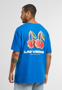 Mėlyna medvilninė marškinėliai su didelėmis vyšnių grafikomis ir užrašu "LAS VEGAS" ant nugaros. Trumpomis rankovėmis ir laisvu siluetu.