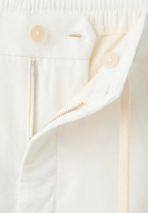 Pantalon écru avec une fermeture éclair ouverte, deux boutons visibles et une ceinture partiellement repliée.