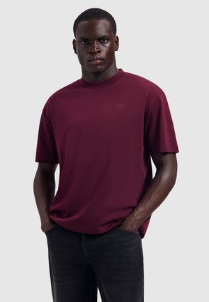 Man wearing a dark red short-sleeve shirt with hands in the pockets of black pants against a plain light background.