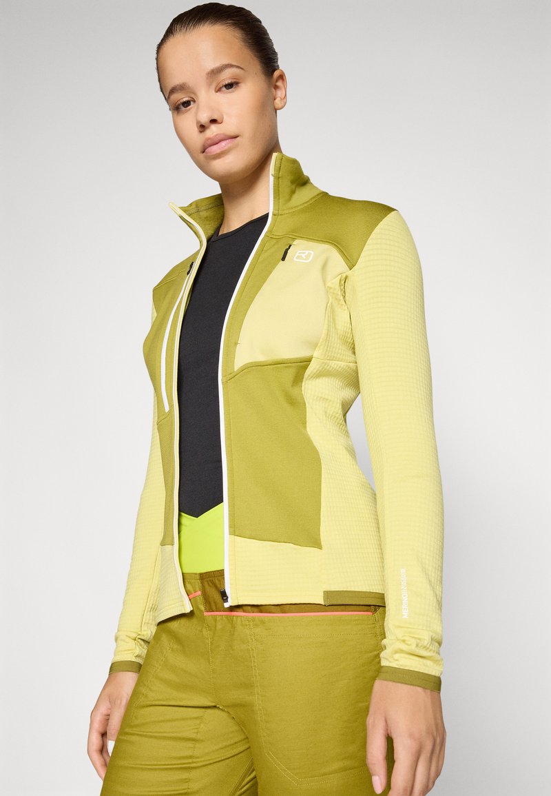Hellgrüne Zip-Jacke mit strukturierten Einsätzen, hohem Kragen und schwarzen Akzenten; kombiniert mit passenden grünen Hosen und einem schwarzen Oberteil darunter.