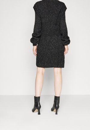 Personne portant une robe en maille noire texturée et des bottines noires à talons, debout, dos à nous, sur un fond clair uni.