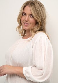 Hvid bluse med tekstureret stof, der har små forhøjede prikker, ballonærmer og en rund halsudskæring, der blødt falder omkring kroppen.