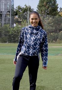 Windbreaker jacket in navy with a geometric white pattern, paired with black athletic pants. Displaying a fitted silhouette and hood.