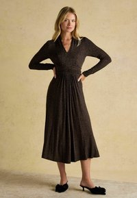 Schwarzes langärmeliges Kleid mit V-Ausschnitt und Tiermuster; hat eine geraffte Taille und einen fließenden Rock. Kombiniert mit schwarzen Mules mit einer Schleifenakzent.