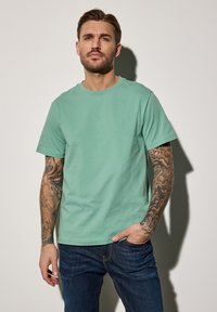 Camiseta de algodón verde menta con escote redondo y mangas cortas, exhibida con vaqueros de denim azul. Presenta una textura suave y un corte relajado.