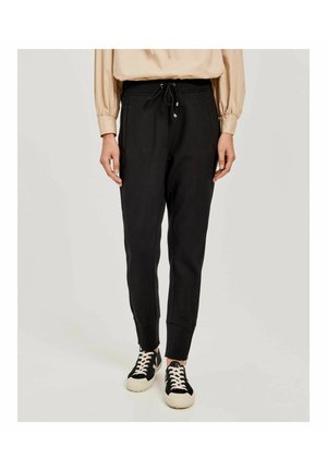 Osoba ubrana w czarne spodnie dresowe z bocznymi kieszeniami oraz czarno-białe trampki, stojąca na neutralnym tle.