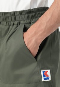 Hånd hviler inne i lommen på olivengrønne shorts med elastisk midje, med en K-Way-logo-patch nær kanten.