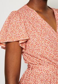 Robe à motif floral avec manches courtes volantées, col en V et taille cintrée sur le haut du torse et le bras d'une personne à la peau foncée.