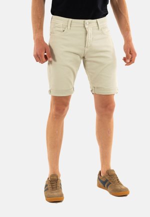 Pantalones cortos de mezclilla color beige claro con puños enrollados, que cuentan con un diseño clásico de cinco bolsillos y cierre de botón metálico. Combinados con zapatillas marrones y azul marino.