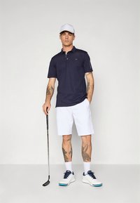 Tmavě modrá polokošile s krátkými rukávy a límečkem. Bílé kraťasy a bílé boty s modrými detaily. Golfová hůl držená v jedné ruce.