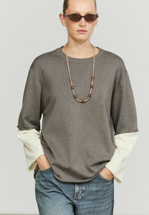 Frau mit brauner Sonnenbrille, geschichtetem grauem und weißem Langarmshirt, blauen Jeans und einer Holzperlenkette mit den Händen in den Taschen.