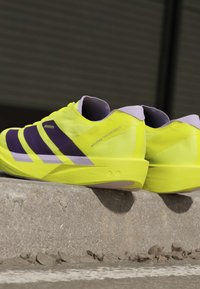 Κίτρινα και μωβ παπούτσια τρεξίματος Adidas Adizero Takumi Sen 11 που ακουμπούν σε μια τσιμεντένια άκρη με θολό σκοτεινό φόντο.