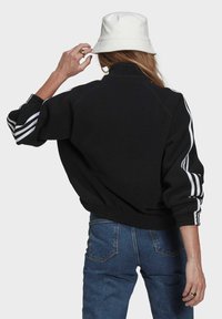 Černá fleecová bunda s vysokým límcem a bílými pruhy na rukávech; spárována s modrými džínami a bílým kloboukem.