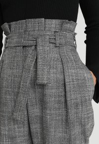 Pantaloni a vita alta a quadri grigi realizzati in un tessuto testurizzato, con vita con cinturino e tasche laterali. Design aderente con pieghe.