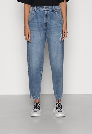 Persoon draagt lichtblauwe high-waisted jeans met lichte slijtage en rafelige zomen, gecombineerd met witte en zwarte chunky sneakers.