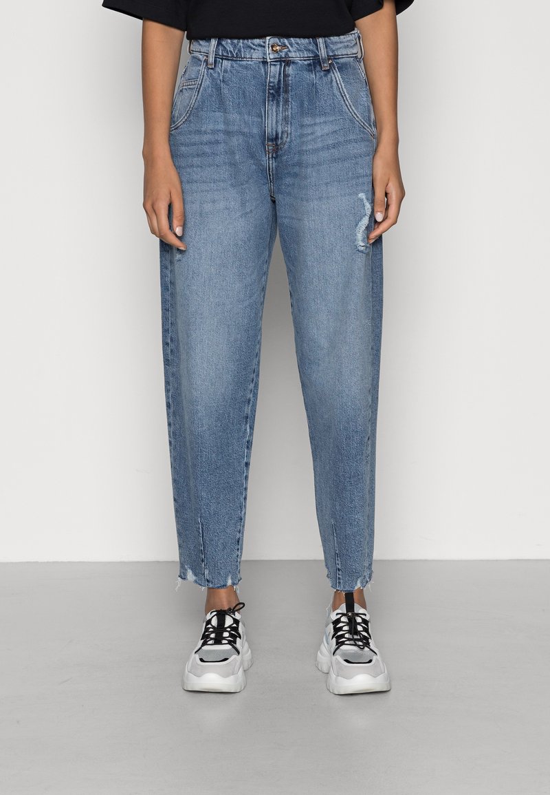 Personne portant un jean taille haute bleu clair avec une légère usure et des ourlets effilochés, associé à des baskets épaisses blanches et noires.