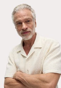 Chemise en tricot à manches courtes, de couleur crème, avec un tissage lâche et un col. Présente deux boutons et une coupe décontractée. Bras croisés devant.