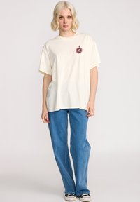T-shirt surdimensionné crème avec un motif floral sur la poitrine gauche, associé à un jean droit bleu clair et des baskets noires.