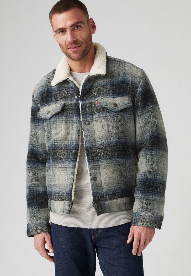 Veste à carreaux en tissu épais, présentant des tons bleus et gris, avec un col en fourrure, fermeture à boutons et poches poitrine. Apparence texturée.