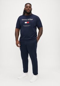 Granatowa koszulka Tommy Hilfiger z białym tekstem logo, zestawiona z dopasowanymi granatowymi dresami i białymi sneakersami, stojąca na jednolitym tle.