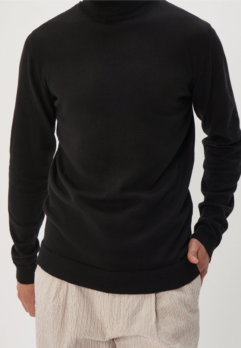 Pull noir à manches longues avec un col montant, fabriqué en un tissu tricot doux. Présente des poignets et un ourlet côtelés, porté sur un pantalon crème à rayures.