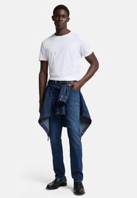 Hvit crew-neck t-skjorte, mørkeblå jeans og svarte skinnsko. En denimjakke bundet rundt livet har detaljer i gullmetall.