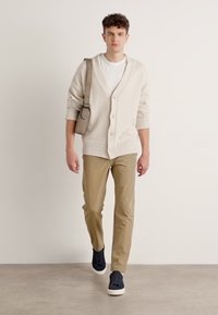 Cardigan bej cu textură nervată, tricou alb, pantaloni khaki și adidași bleumarin. Accesoriile includ o geantă tip crossbody.
