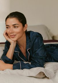 Navy floral-patterned satin pajama set with pink piping, featuring long sleeves and relaxed fit, laid against light-colored bedding.