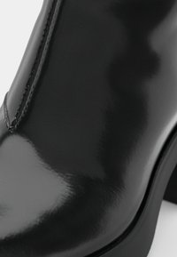 Černá kožená kotníková bota s lesklým povrchem, bočním zipem a masivním podpatkem. Hladká textura a minimalistické designové detaily jsou výrazné.
