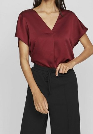 Vrouw draagt een bordeauxrode V-hals blouse met korte mouwen, ingestopt in een zwarte hoge taille broek met een trekkoord in de taille, poseert met hand op de heup.