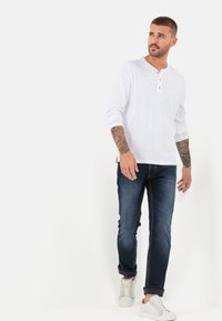 Biała, długorękawowa koszula henley z dekoltem zapinanym na trzy guziki, w połączeniu z ciemnoniebieskimi dżinsami i białymi trampkami. Gładka tekstura i luźny krój.
