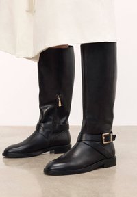 Fekete bőr, térd felett érő csizmák sima textúrával, oldalsó cipzárral és bucklis pánttal. Lapos talpakkal és minimalista dizájnnal.