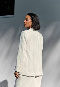 Blazer blanc à rayures avec fermeture à bouton unique, coupe ajustée et tissu texturé. Il présente des revers nets et une fente au dos.