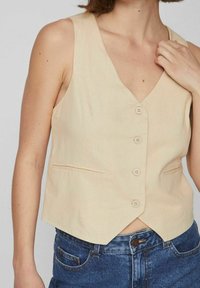 Béžová lněná vesta s hlubokým výstřihem do V, pěti knoflíky a bočními kapsami. Hladká textura, přiléhavý střih, kombinovaná s modrými džínovými šortkami.