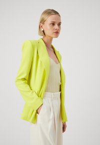 Sacou verde lime cu umeri croiți, material neted și buzunare frontale, asortat cu un top bej deschis cu benzi și pantaloni crem.