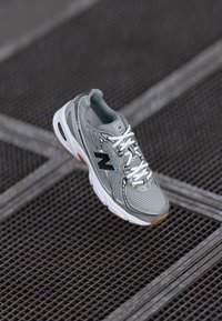 Scarpa da corsa New Balance grigia con lacci bianchi e suola in gomma appoggiata su una superficie metallica a griglia inclinata diagonalmente.