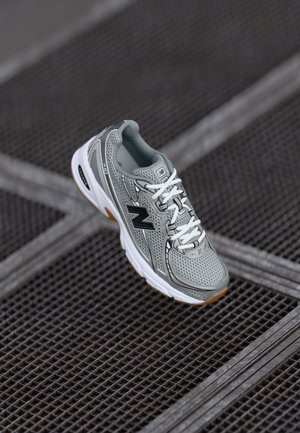 Grijze New Balance hardloopschoen met witte veters en gumzool, rustend op een metalen roosteroppervlak in een diagonale hoek.