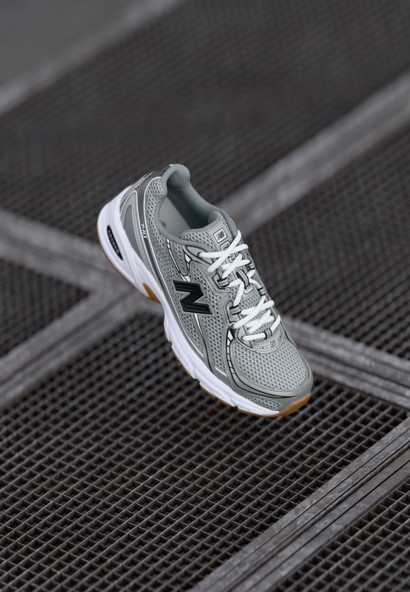Scarpa da corsa New Balance grigia con lacci bianchi e suola in gomma appoggiata su una superficie metallica a griglia inclinata diagonalmente.