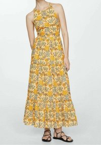 Abito lungo giallo con motivi floreali e foglie, caratterizzato da un corpetto aderente, balze a strati e scollo all'americana. Abbinato a sandali neri.