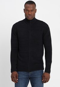 Svart zip-up tröja med ribbat mönster och hög krage, med en figurnära design och långa ärmar. Bärs över blå jeans.