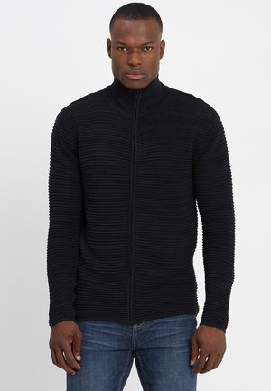 Svart zip-up genser med ribbet tekstur og høyt krage, med en tettsittende design og lange ermer. Brukt over blå jeans.