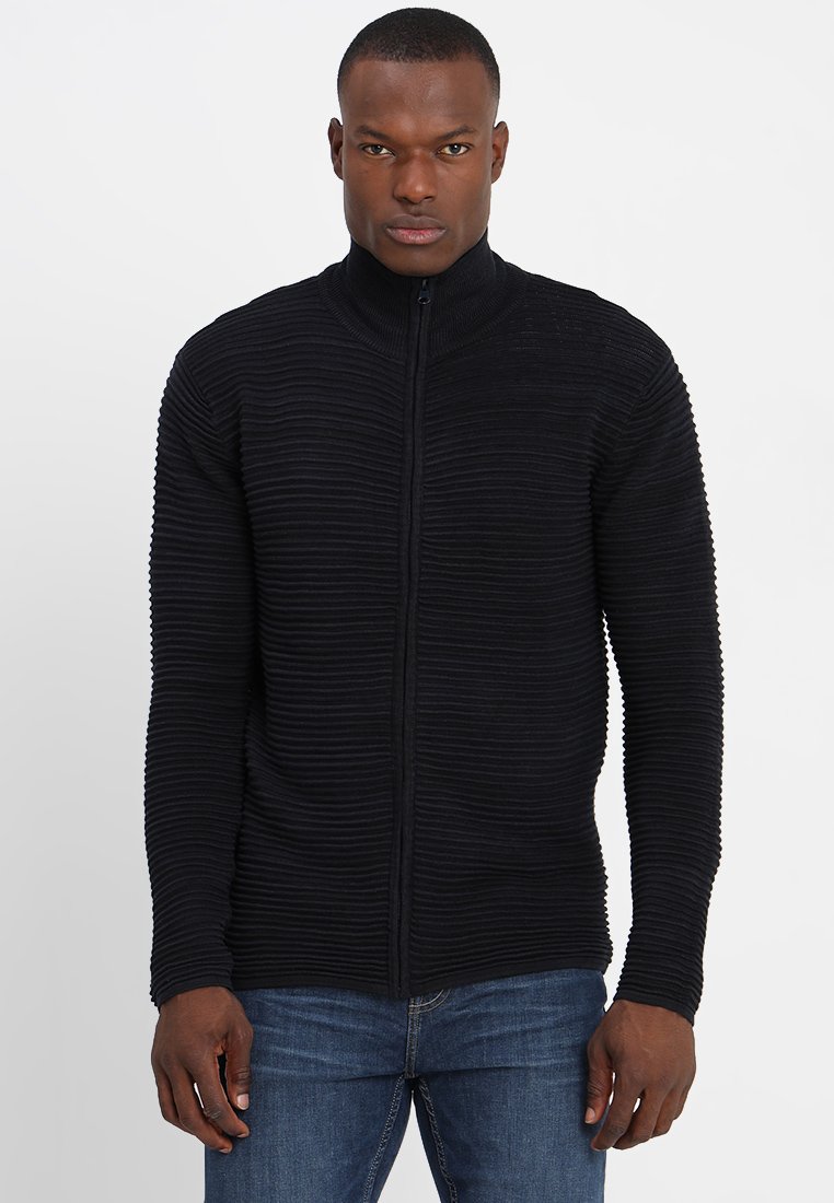 Svart zip-up tröja med ribbat mönster och hög krage, med en figurnära design och långa ärmar. Bärs över blå jeans.