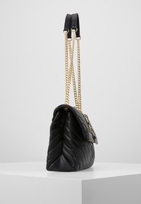 Černá prošívaná kožená kabelka se zlatými řetízkovými popruhy a černými koženými ramenními podložkami, vystavená na bílé podstavě proti šedému pozadí.