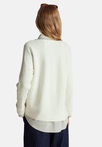 Kremowy sweter o prążkowanej fakturze, z kontrastowym paskiem u dołu i długimi rękawami. Tył ma luźny krój.